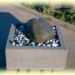 Dunkler       Granit- Quellstein auf hellgrauem Zierkies in Basalt-Becken (Größe       80/80/35 cm)
